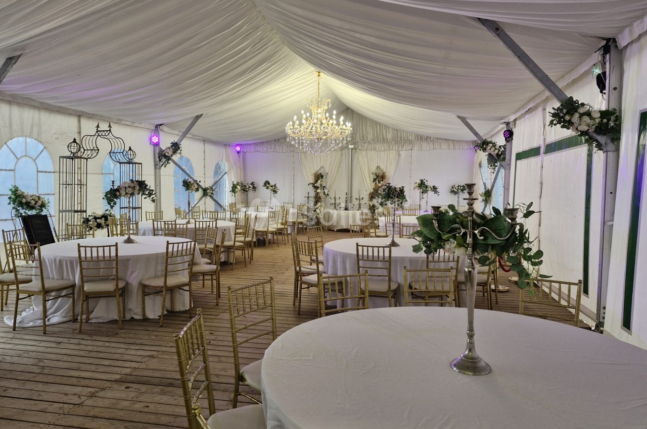 Intérieur de chapiteau drapé blanc avec tables rondes napées, chaises dorées, lustre et décoration florale.