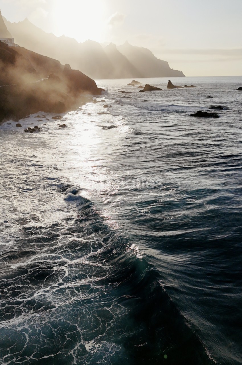 Côte rocheuse avec vagues déferlantes sous un ciel brumeux et falaises en arrière-plan.