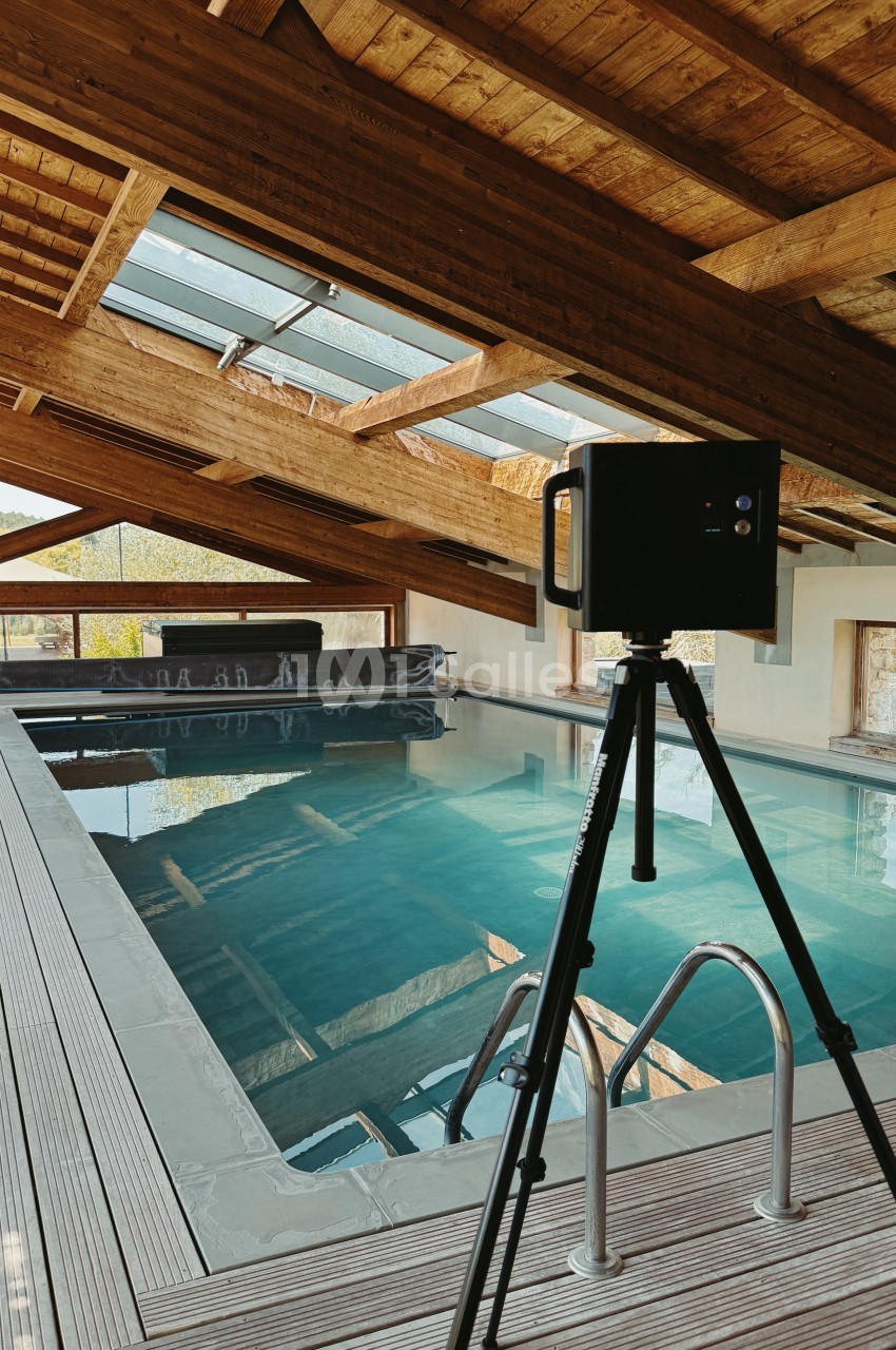 Piscine intérieure aux eaux turquoises avec charpente bois apparente et vasistas - caméra sur trépied au premier plan.