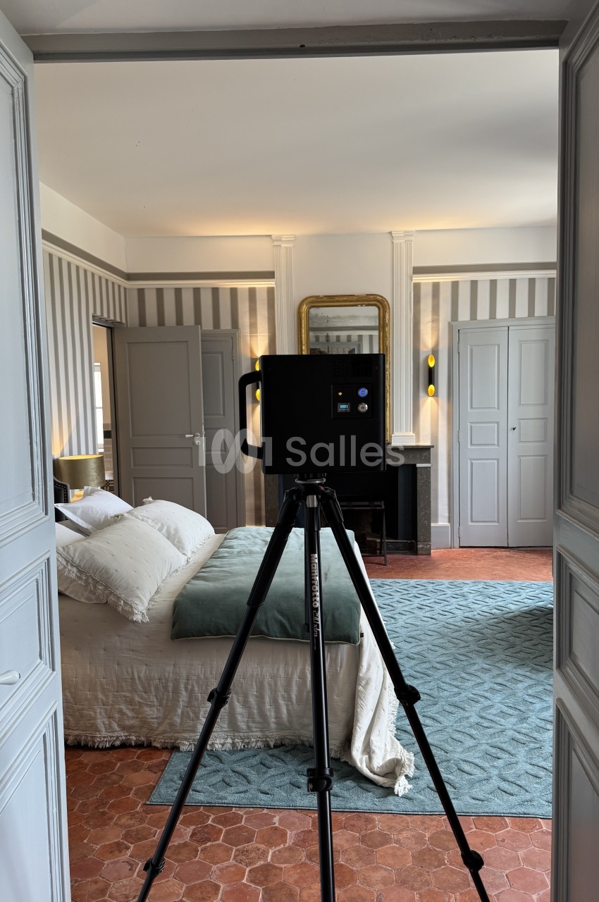 Chambre classique avec lit, colonnes blanches, miroir doré et camera vidéo sur trépied.
