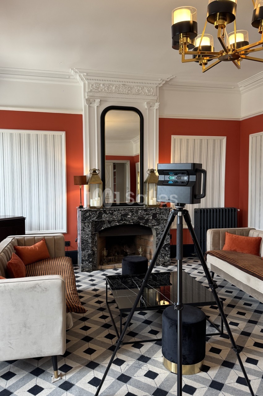 Intérieur avec murs rouges, cheminée en marbre noir, miroir encadré blanc, canapés beige et équipement photographique sur trépied.