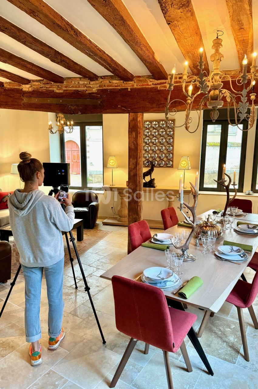 Salle avec poutres apparentes, table dressée avec chaises burgundy et chandelier doré, personne photographiant.