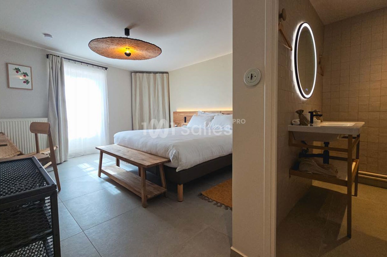 Chambre double avec lit blanc, salle de bain attenante, luminaire suspendu doré et fenêtre sur terrasse.