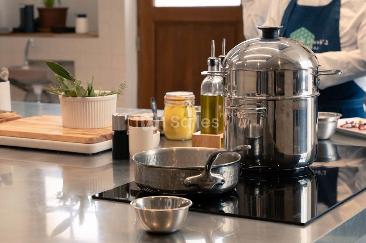 Cuisine moderne avec plan de travail gris, ustensiles en inox, pot avec couvercle, poêle, bocaux d'huile et plante verte.