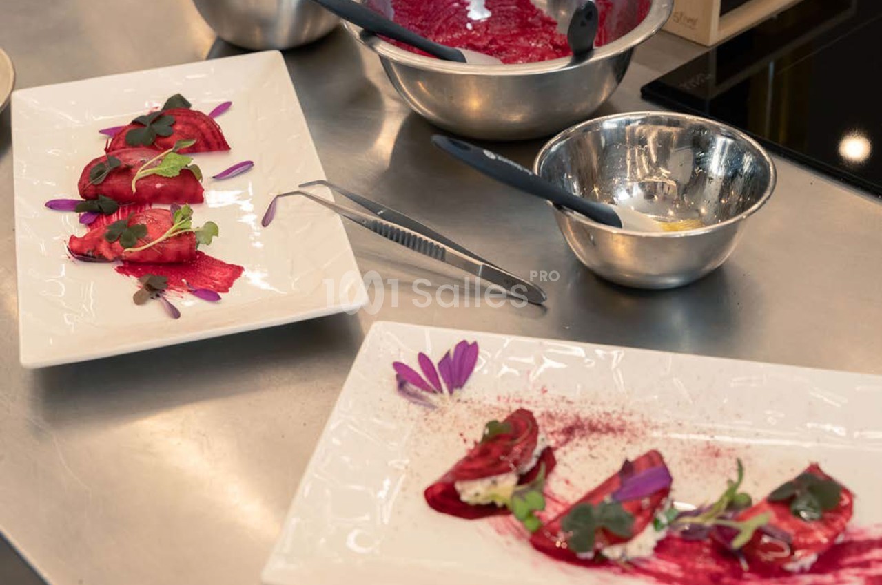 Plan de travail de cuisine avec deux assiettes de dressage de plats à base de fraises rouges et fleurs violettes, ustensiles métalliques.