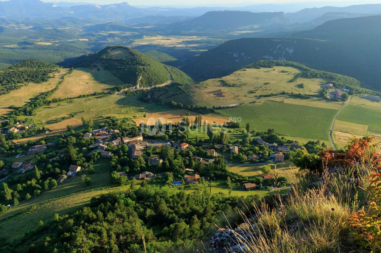 Vallée verdoyante avec village niché entre forêts, prairies et montagnes en arrière-plan sous ciel dégagé.
