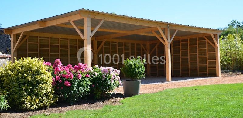 Abri en bois ouvert avec toit en pente, entouré de fleurs, d'arbustes et d'une pelouse verte.