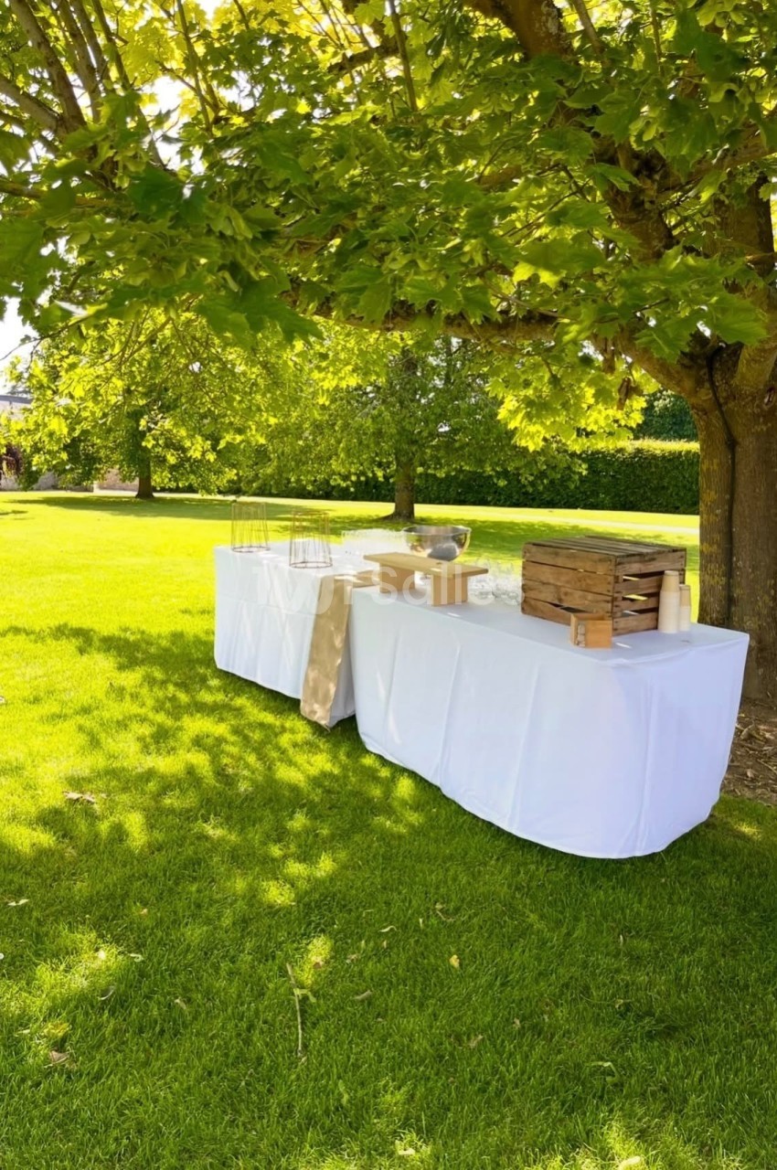 Table dressée sous un arbre dans un jardin, avec nappes blanches et éléments de service en bois et métal.
