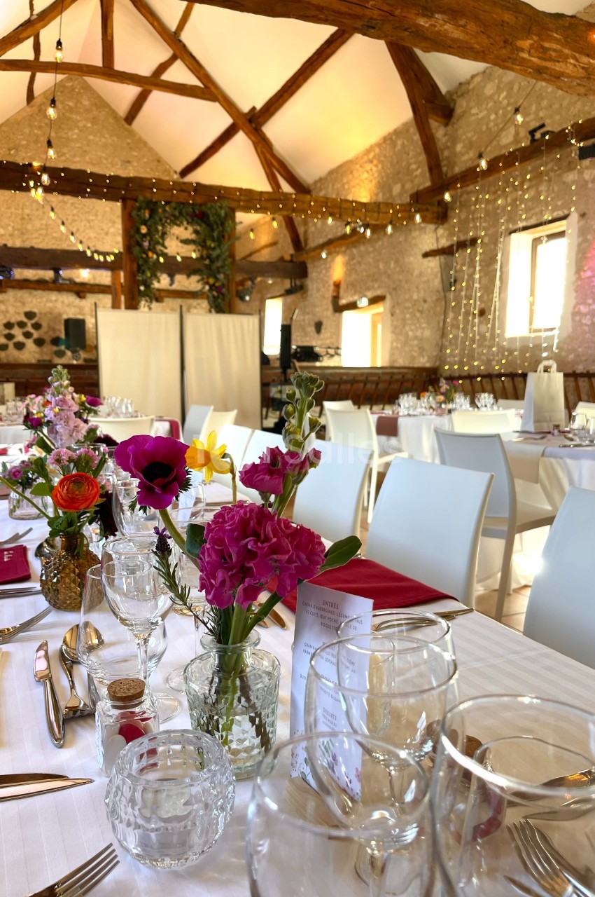 Salle de réception décorée pour un événement, avec tables dressées, fleurs colorées et guirlandes lumineuses suspendues.