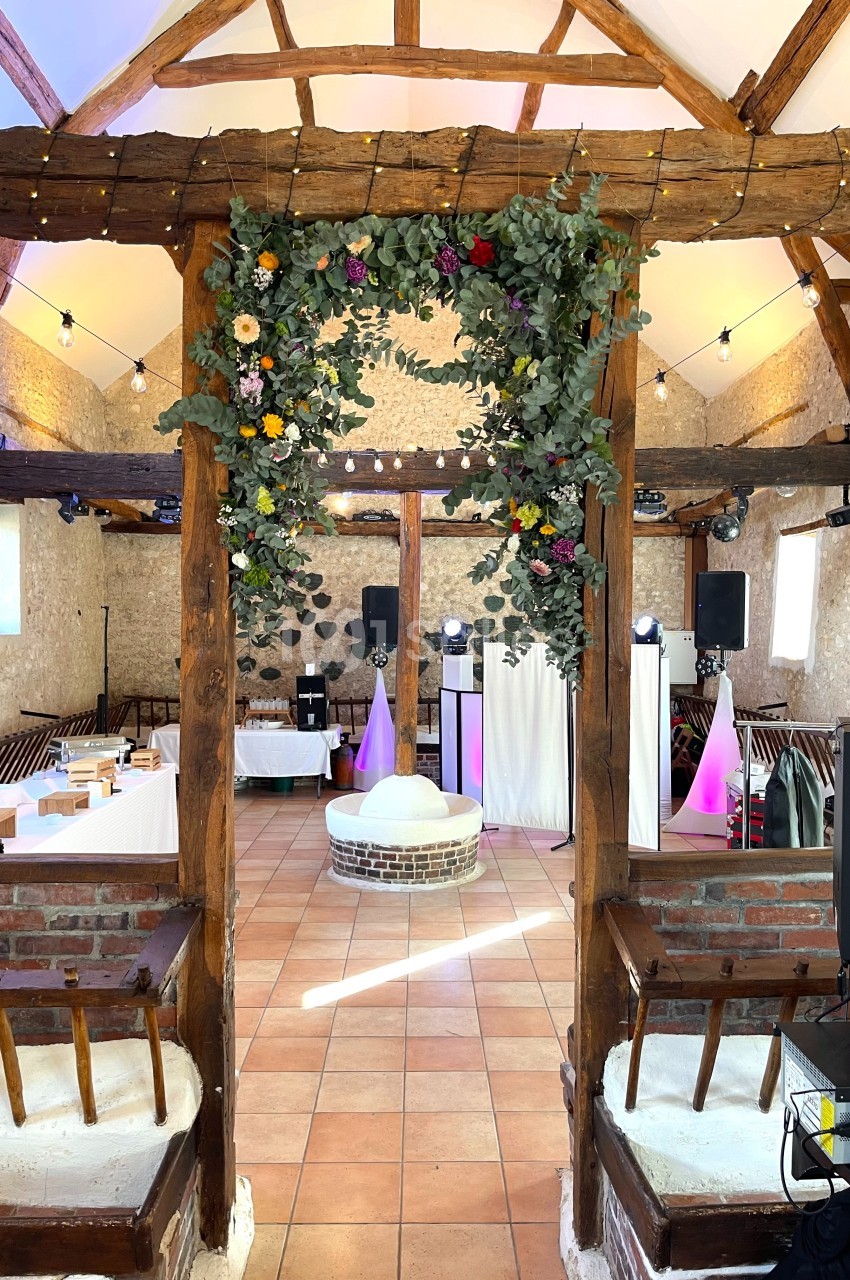Salle de réception rustique avec poutres apparentes, décor floral suspendu et éclairage festif.