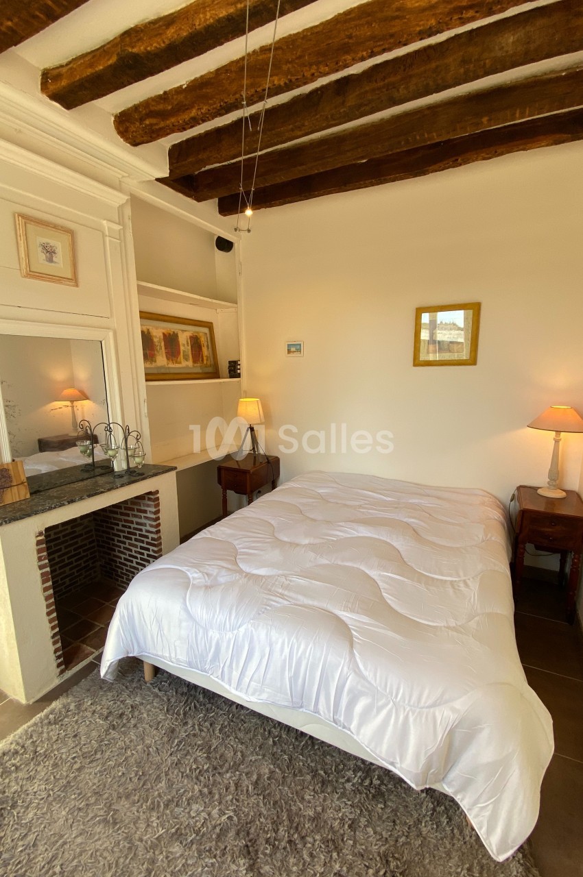Chambre lumineuse avec poutres apparentes, lit double, tables de chevet, lampes et cheminée décorative.