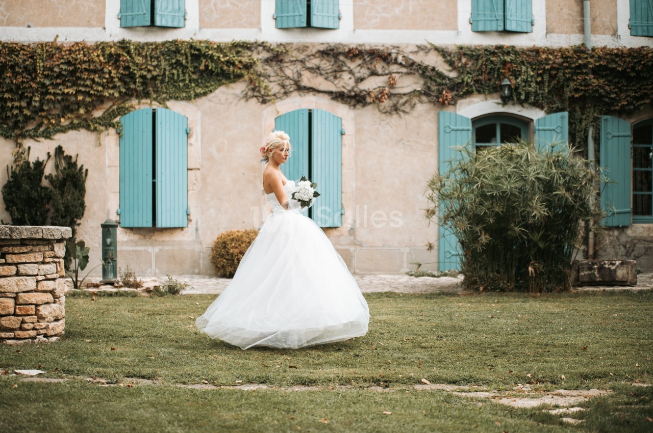 Une mariée en robe blanche tient un bouquet dans un jardin devant une maison aux volets bleus.