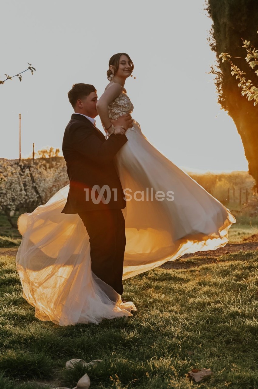 Un homme en costume soulève une femme en robe blanche dans un jardin au coucher du soleil.