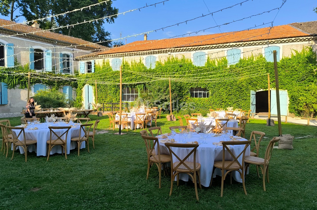 Tables rondes dressées pour un événement en plein air dans une cour verdoyante, entourée de bâtiments anciens.