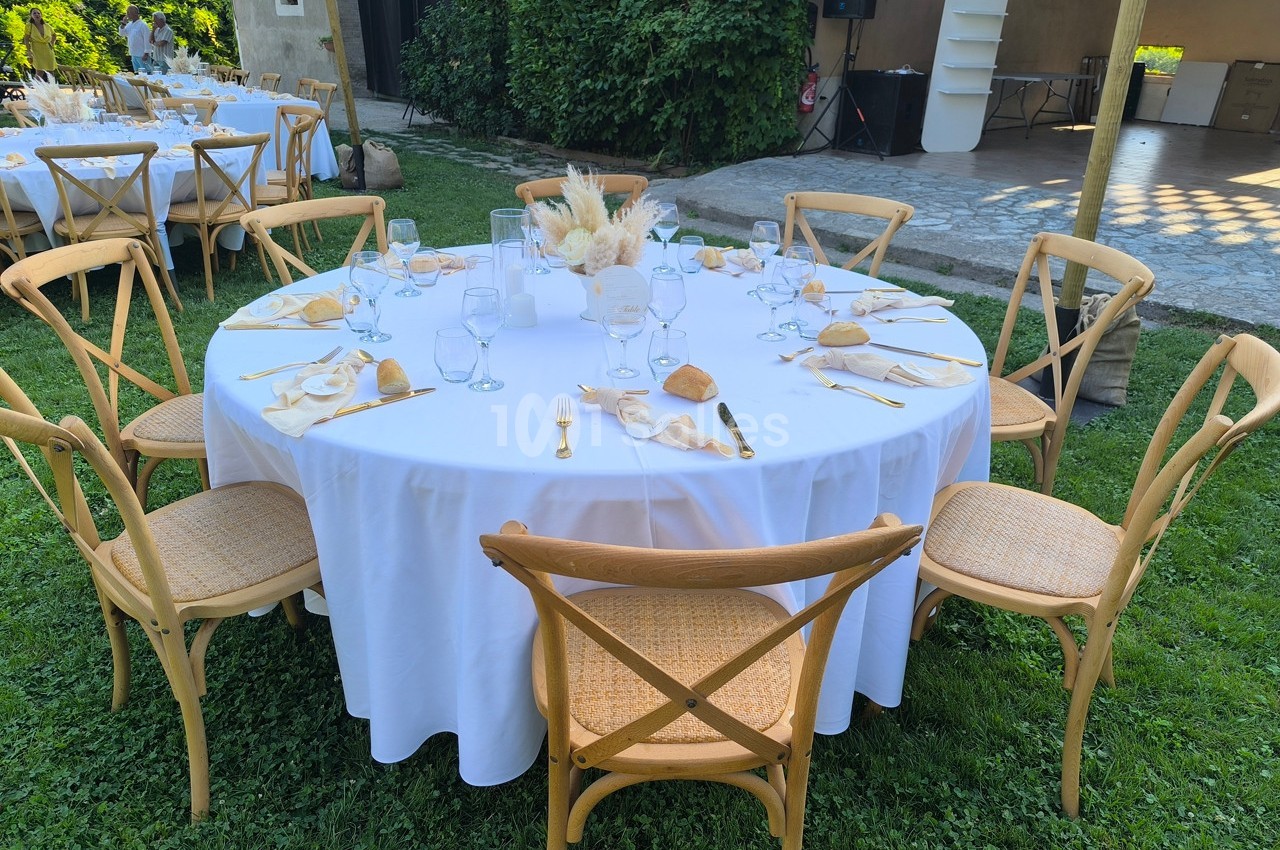 Table ronde dressée pour un repas en extérieur, avec nappes blanches, couverts dorés et décoration florale centrale.