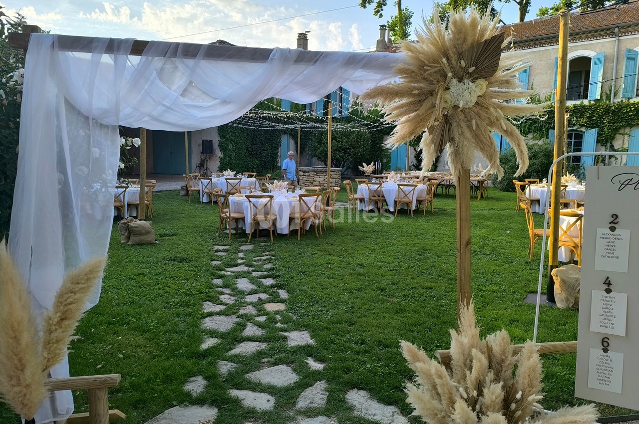 Décoration de mariage en extérieur avec arche ornée de voilages et de pampas, tables dressées sur une pelouse.