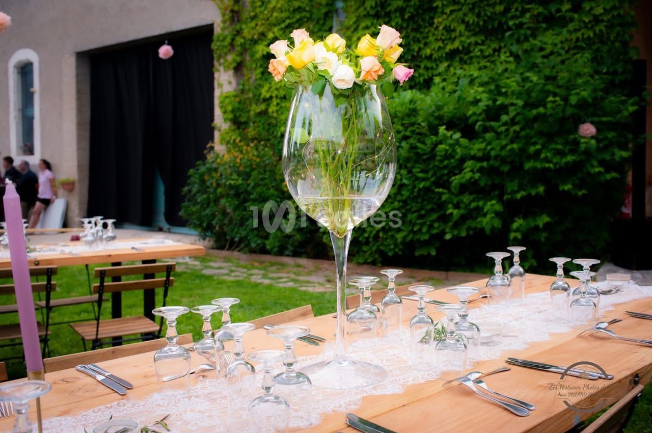 Centre de table avec un grand vase en forme de verre rempli d'eau et de fleurs, sur une table en bois dressée en extérieur.