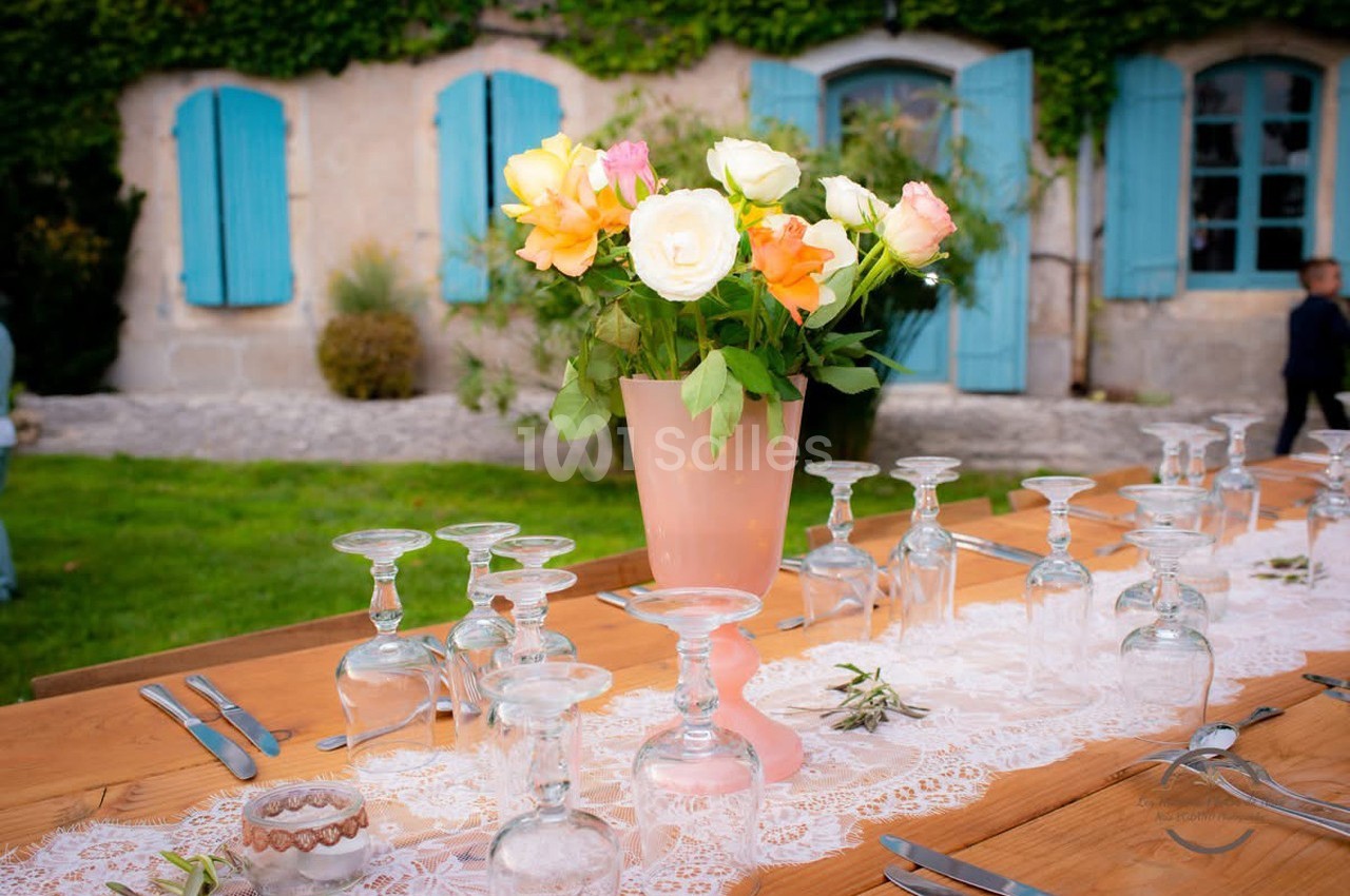 Vase de fleurs colorées sur une table en bois décorée, devant une maison en pierre aux volets bleus.