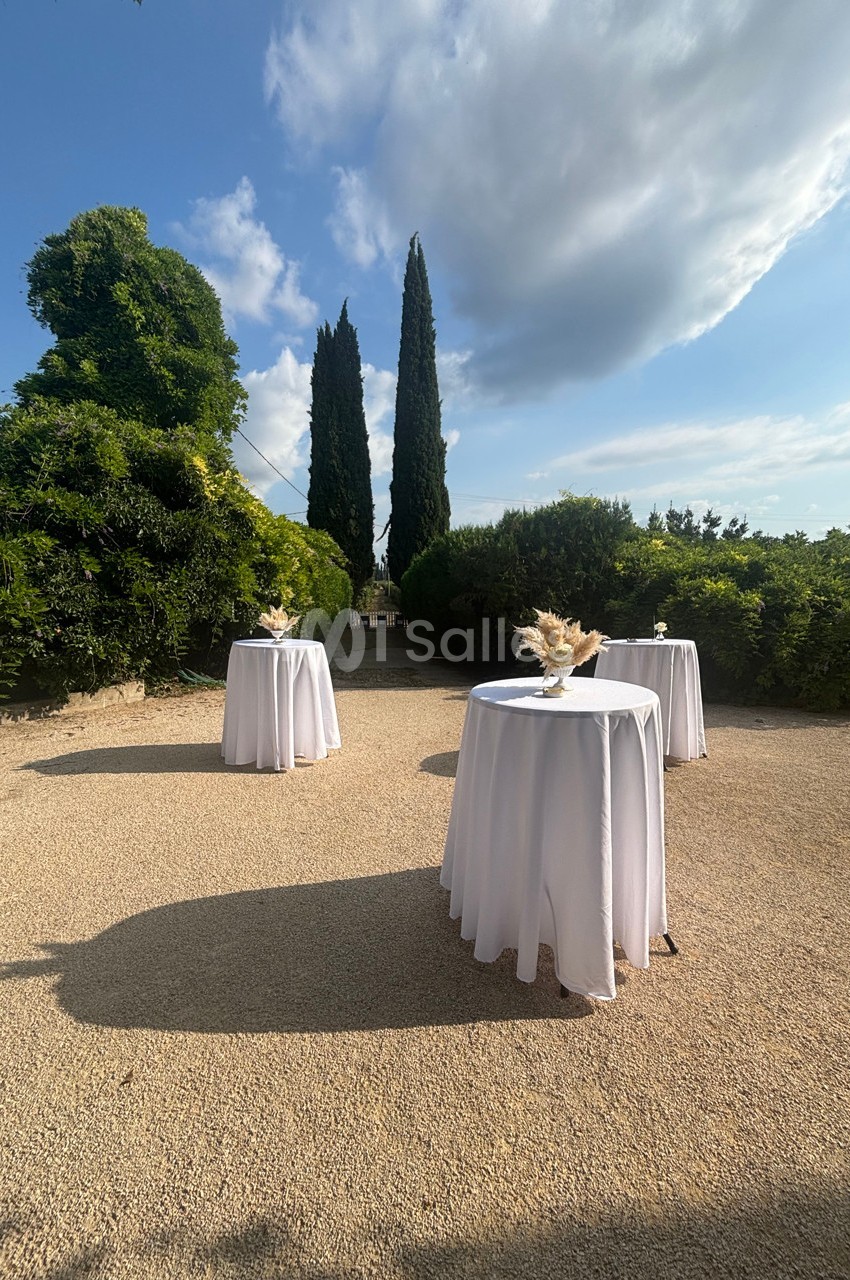 Trois tables hautes drapées de nappes blanches disposées en extérieur, entourées de verdure et de cyprès.