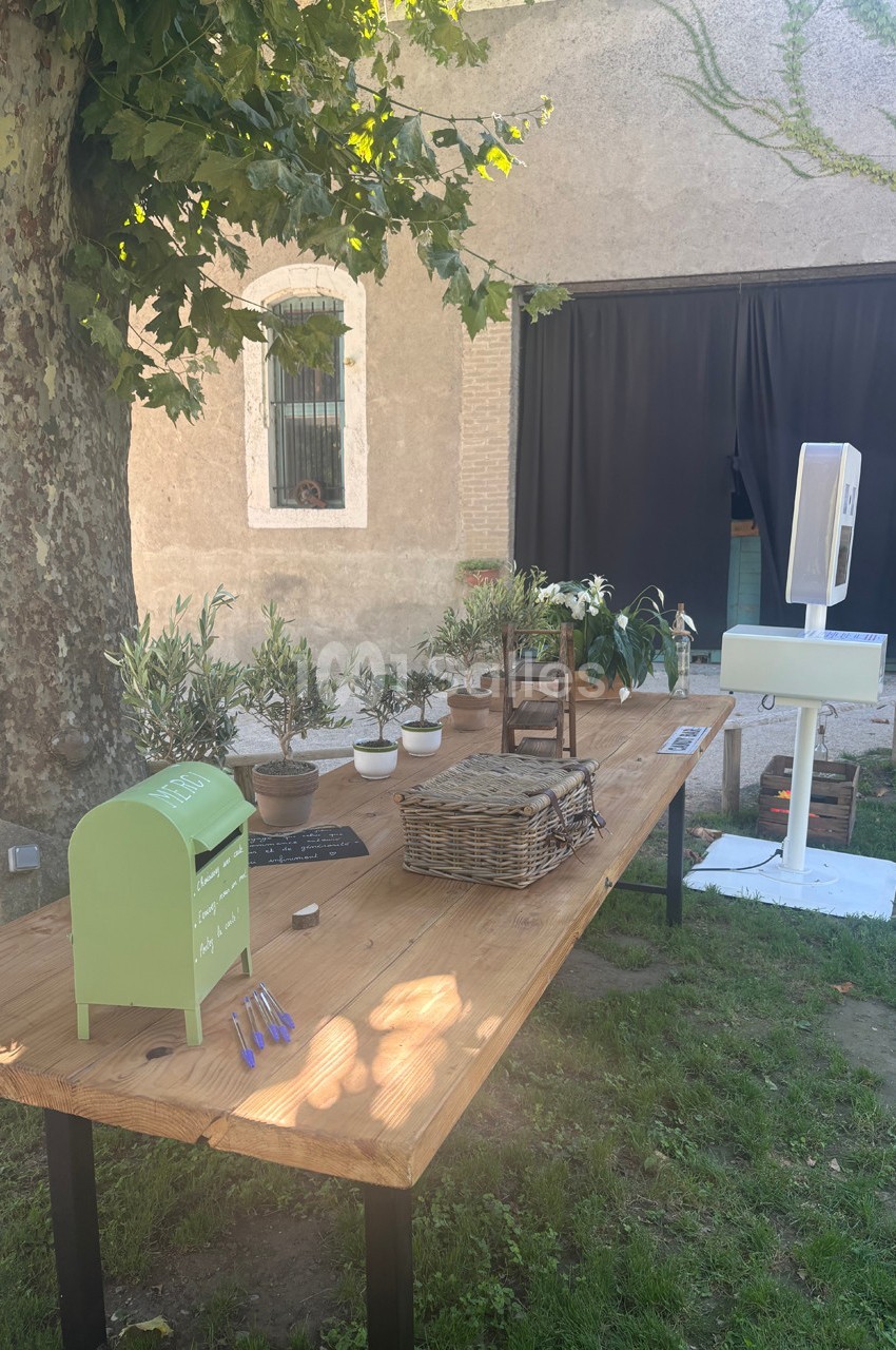 Table en bois à l'extérieur avec des plantes en pot, une boîte aux lettres verte, des paniers et un photomaton en arrière…