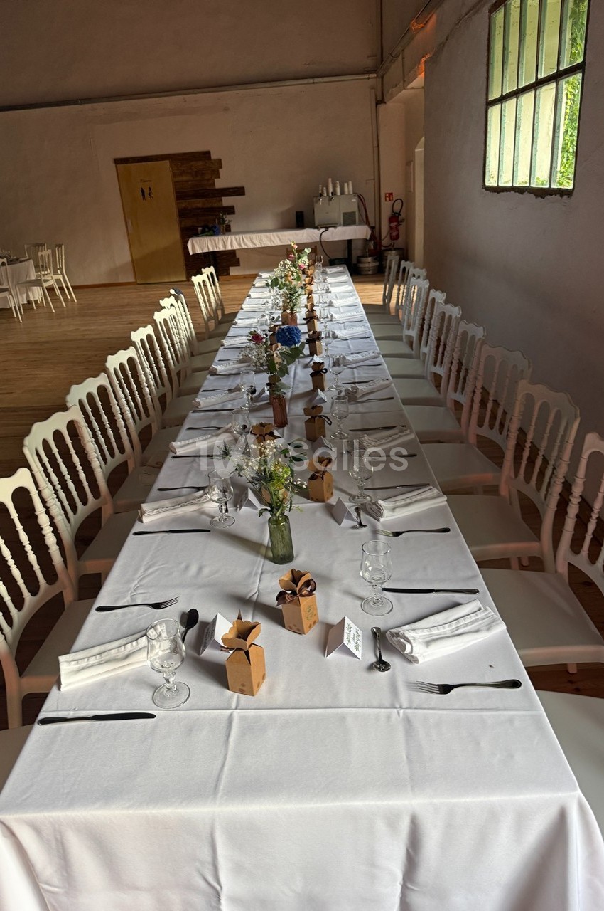 Table longue dressée pour un repas, avec nappes blanches, chaises assorties, décorations florales et petits cadeaux.