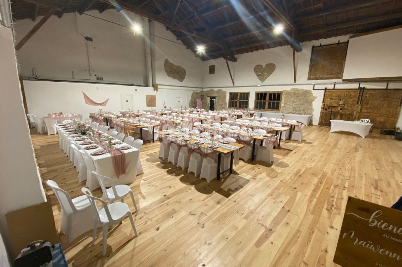 Salle de réception avec tables décorées pour un événement, chaises alignées et plafond en bois apparent.