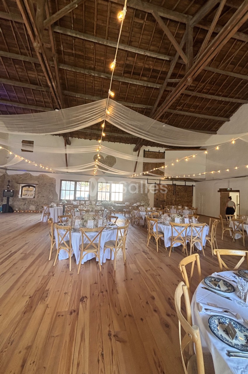 Salle de réception avec tables dressées, chaises en bois, guirlandes lumineuses et plafond en poutres apparentes.