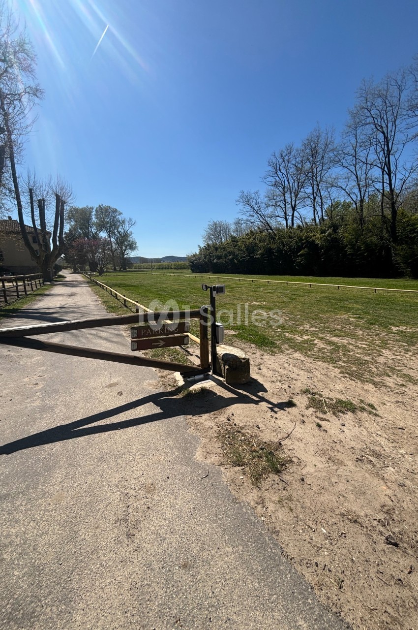 Barrière en bois fermant l'accès à un parking herbeux, entouré d'arbres sous un ciel dégagé.