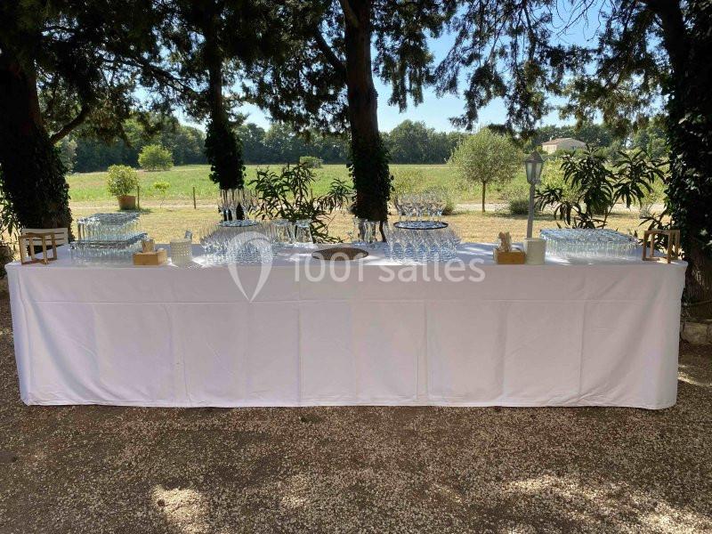 Table dressée en extérieur avec nappage blanc, verres et plateaux, entourée de végétation et éclairée par la lumière…
