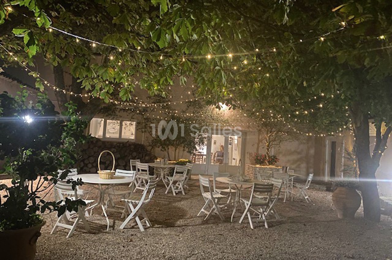 Terrasse extérieure éclairée par des guirlandes lumineuses, avec des tables et chaises blanches sous des arbres.