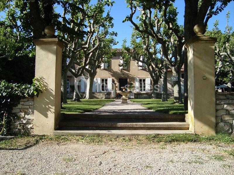 Entrée d'une maison provençale entourée d'arbres, avec une allée menant à une fontaine devant la façade.