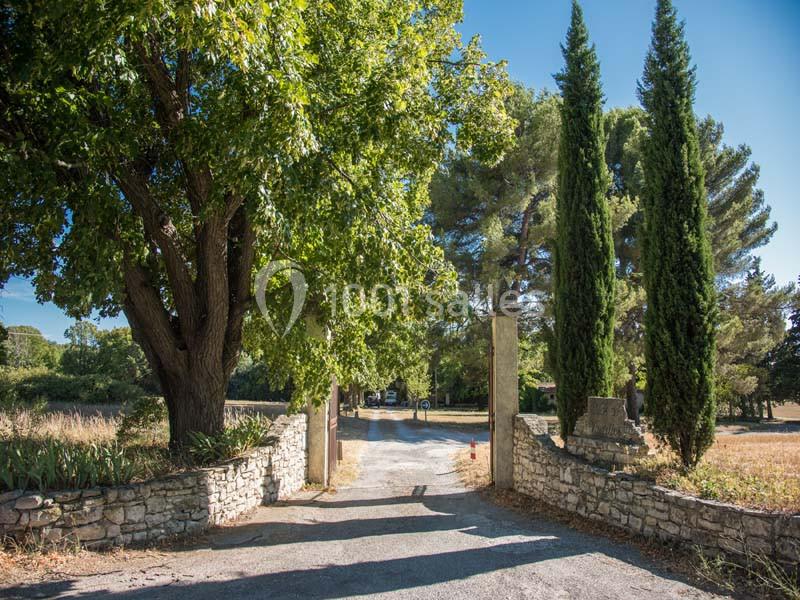 Entrée d'une propriété bordée de murs en pierre, avec des arbres et un chemin menant à un espace dégagé.
