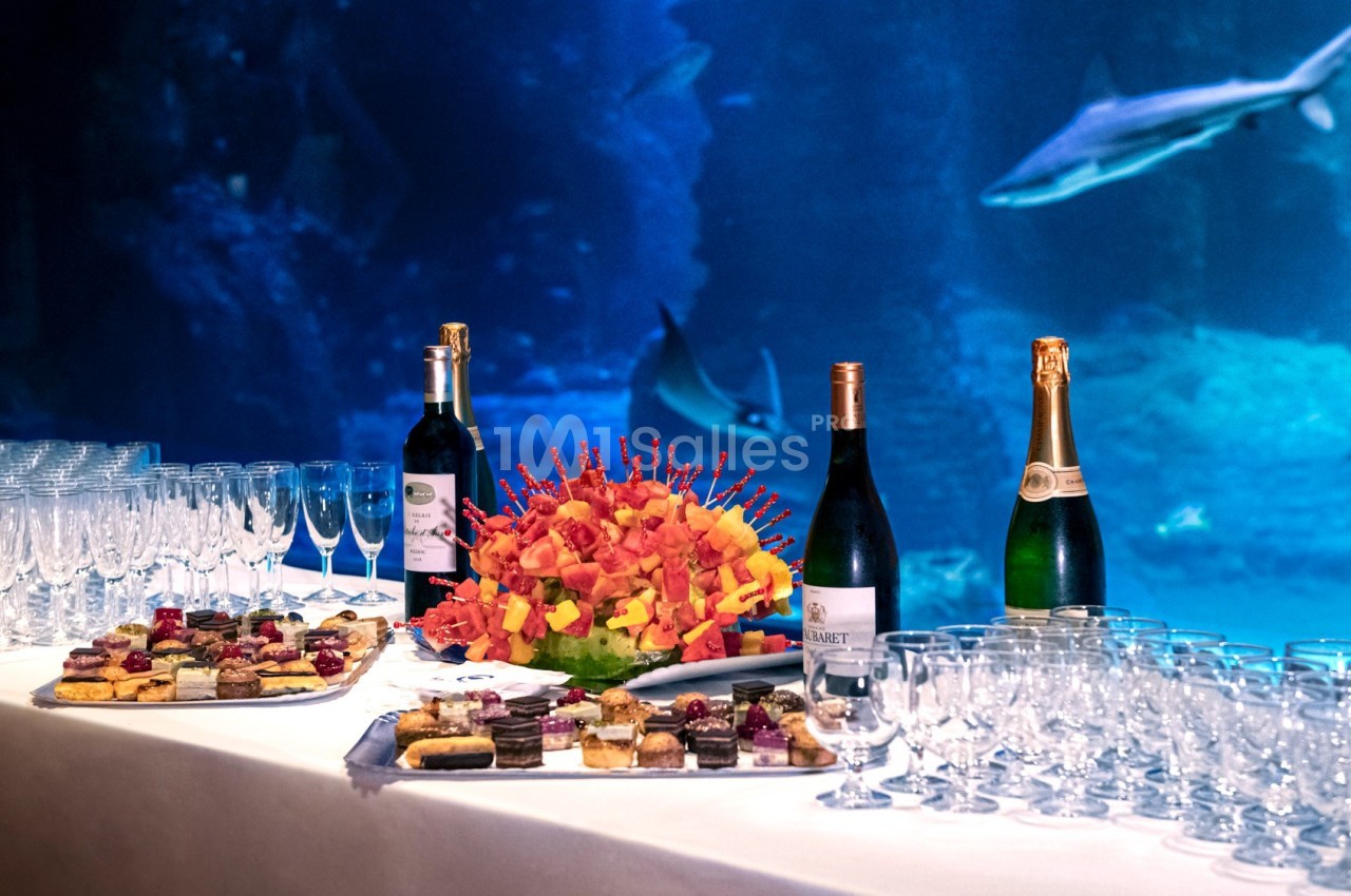 Table dressée avec des verres, bouteilles de vin et champagne, assortiments de desserts, devant un aquarium avec des requins.