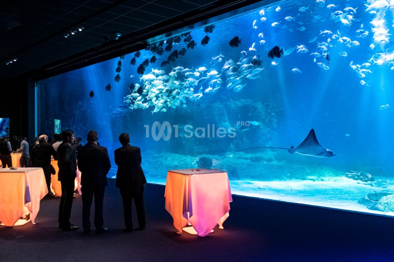 Des personnes debout près de tables éclairées regardent un grand aquarium avec des poissons et une raie nageant.