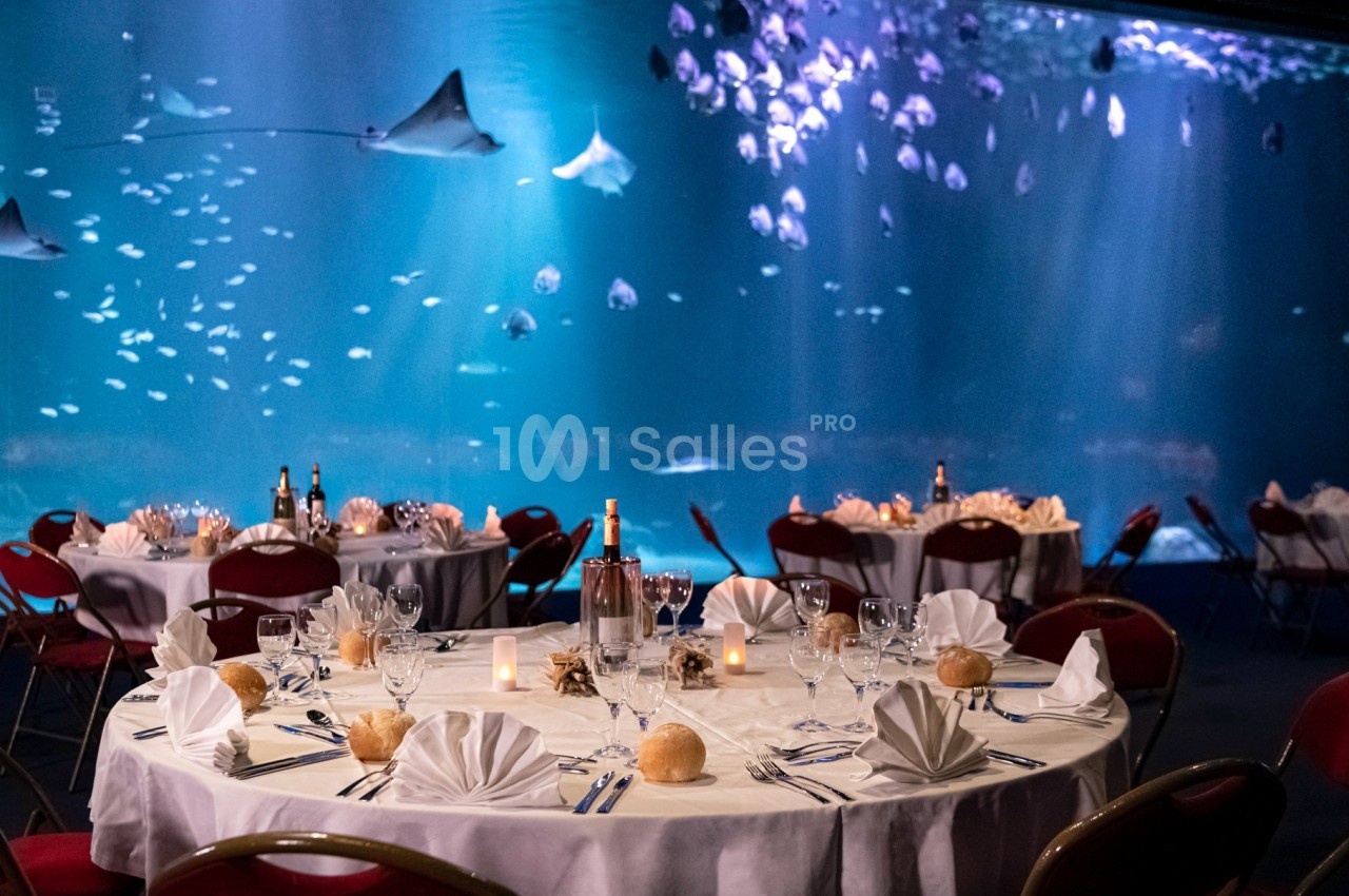 Salle de réception avec tables dressées, nappes blanches et vue sur un grand aquarium rempli de raies et poissons.