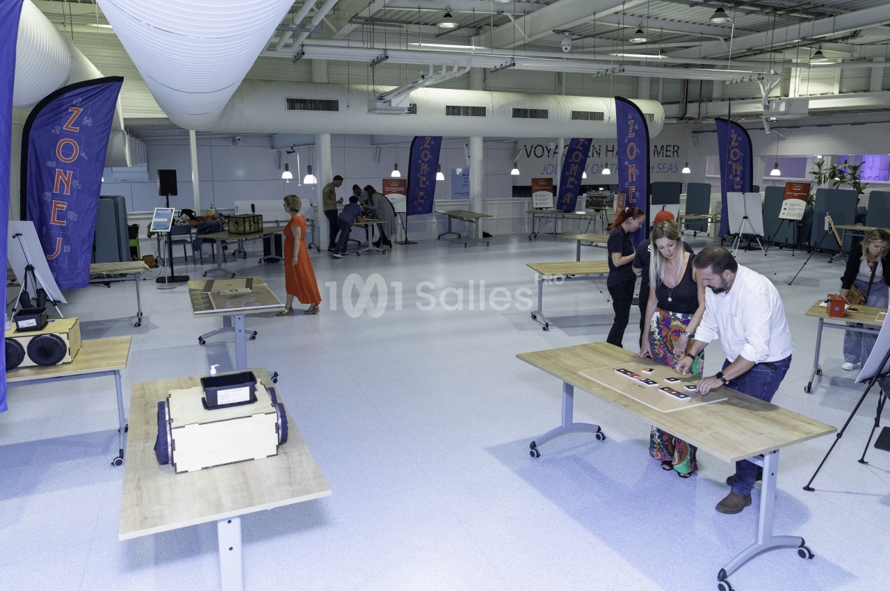 Salle lumineuse avec des tables en bois, des stands d'exposition et des personnes observant ou manipulant des objets.