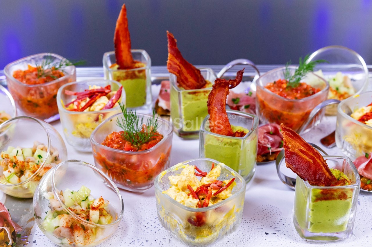 Assortiment de verrines colorées garnies de légumes, sauces, bacon et herbes fraîches, présentées sur une nappe blanche.