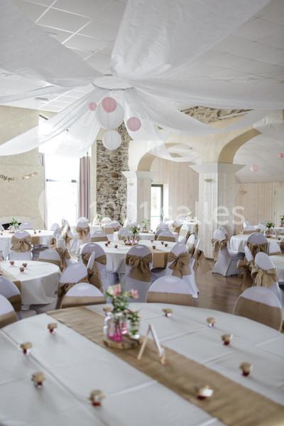 Salle de réception décorée avec des tables rondes, nappes blanches, chaises ornées de nœuds et lanternes suspendues.