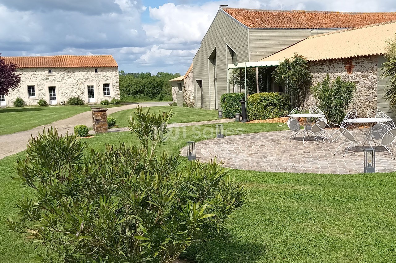 Cour extérieure avec tables et chaises sur une terrasse pavée, entourée de pelouse et bâtiments en pierre sous un ciel…