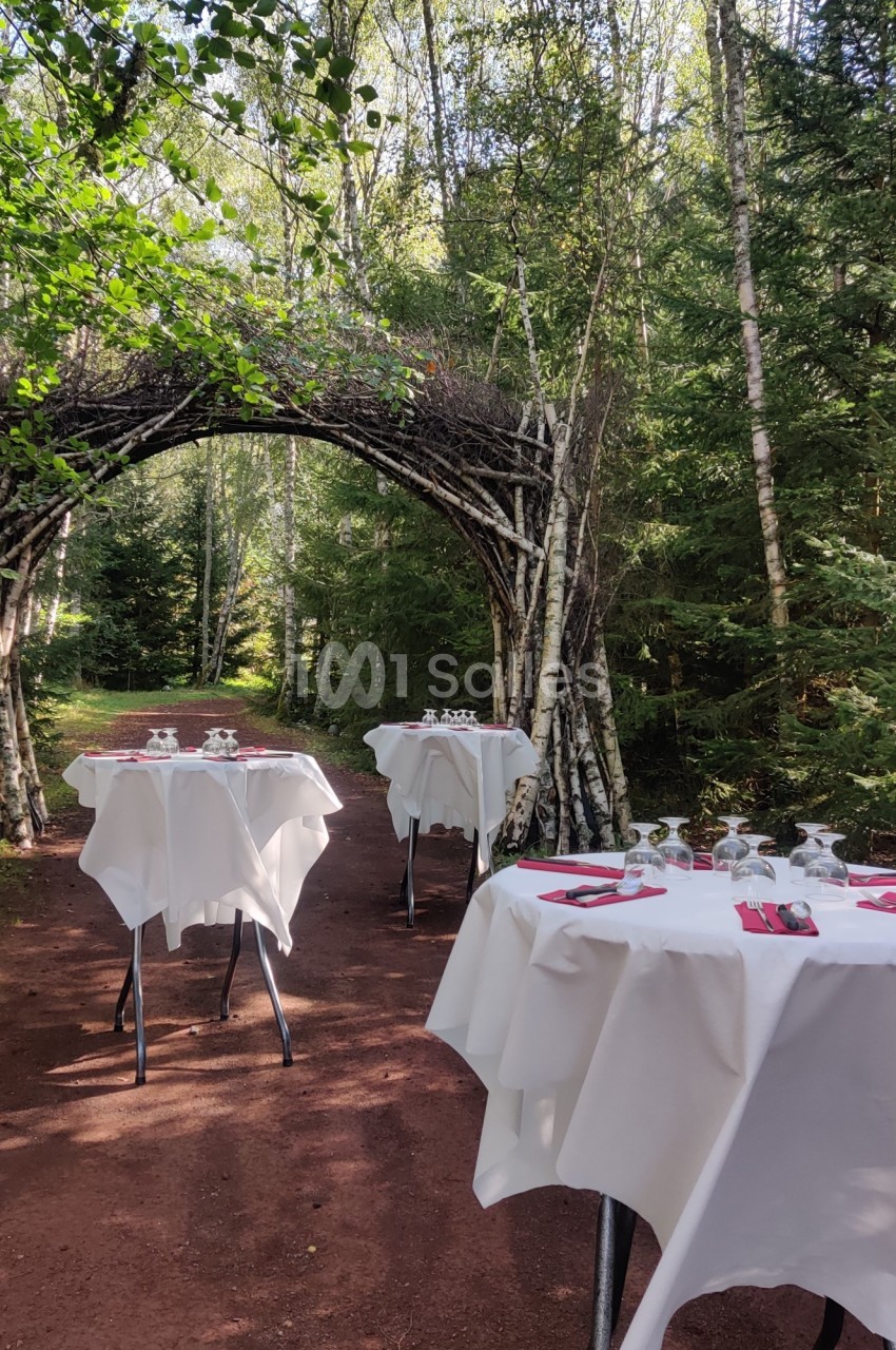 Tables dressées avec nappes blanches et couverts, disposées en extérieur sur un chemin forestier sous une arche en bois.