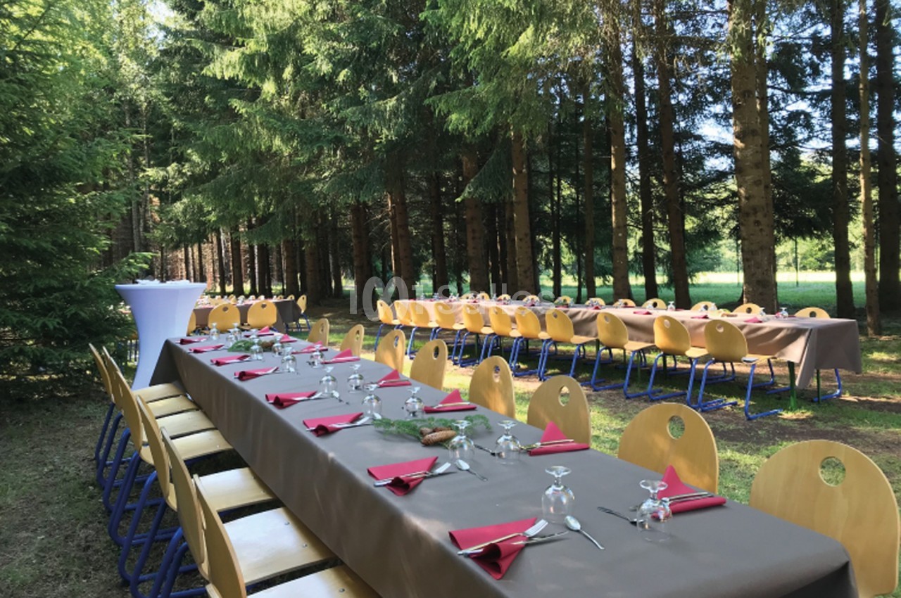 Tables dressées avec nappes brunes, serviettes rouges et verres, disposées en extérieur dans une forêt.