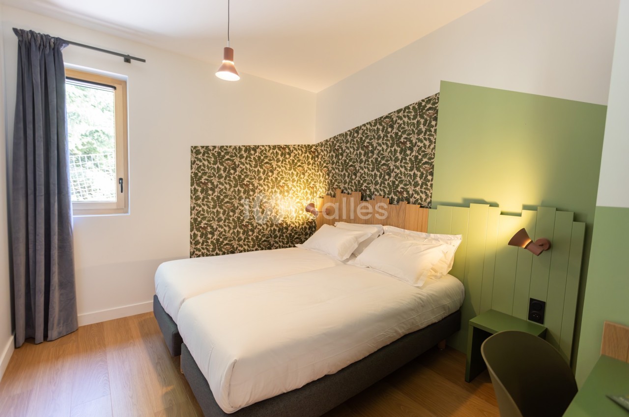Chambre lumineuse avec lit double, mur décoré de papier peint à motifs végétaux et éclairage d'appoint.