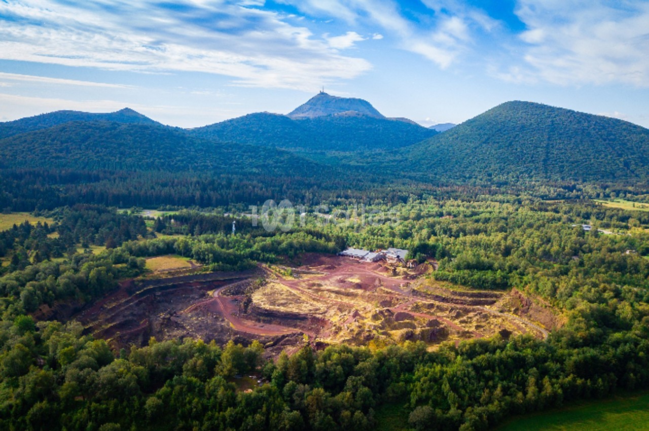 Vue aérienne d'une carrière entourée de forêts avec des montagnes en arrière-plan sous un ciel partiellement nuageux.