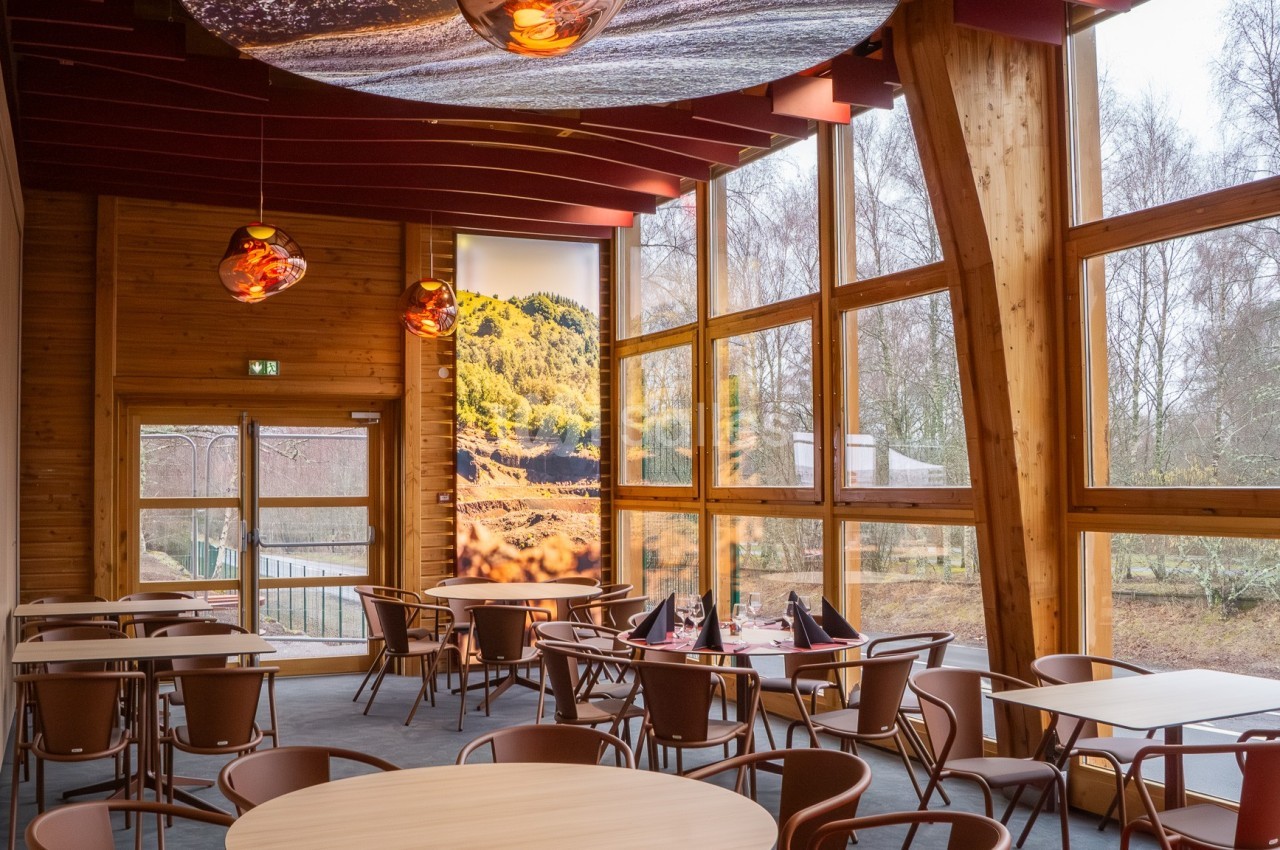 Salle de restaurant lumineuse avec de grandes baies vitrées, des tables en bois et des chaises marron.