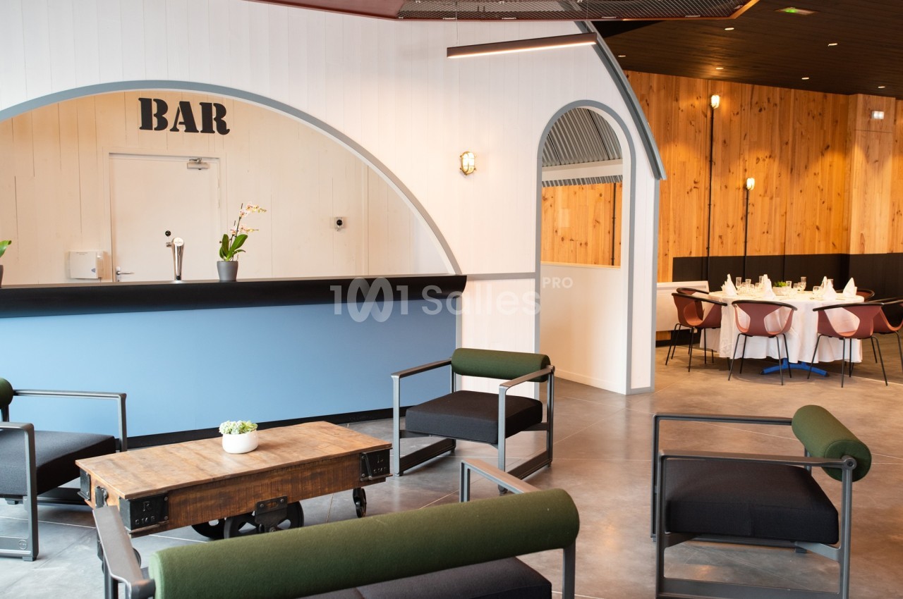 Espace intérieur avec un bar moderne, des fauteuils verts et noirs, et une table en bois, salle à manger en arrière-plan.