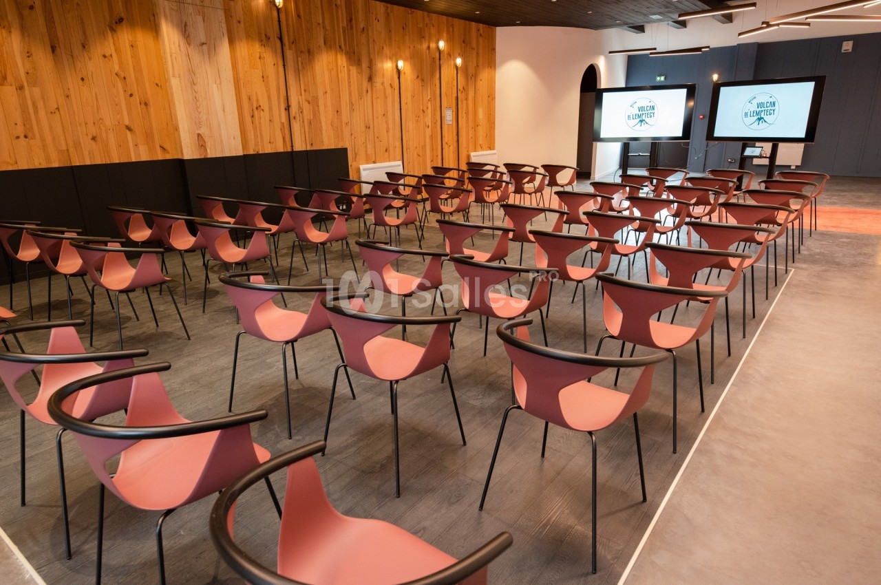 Salle de conférence moderne avec des rangées de chaises rouges et deux écrans affichant un logo à l'arrière-plan.