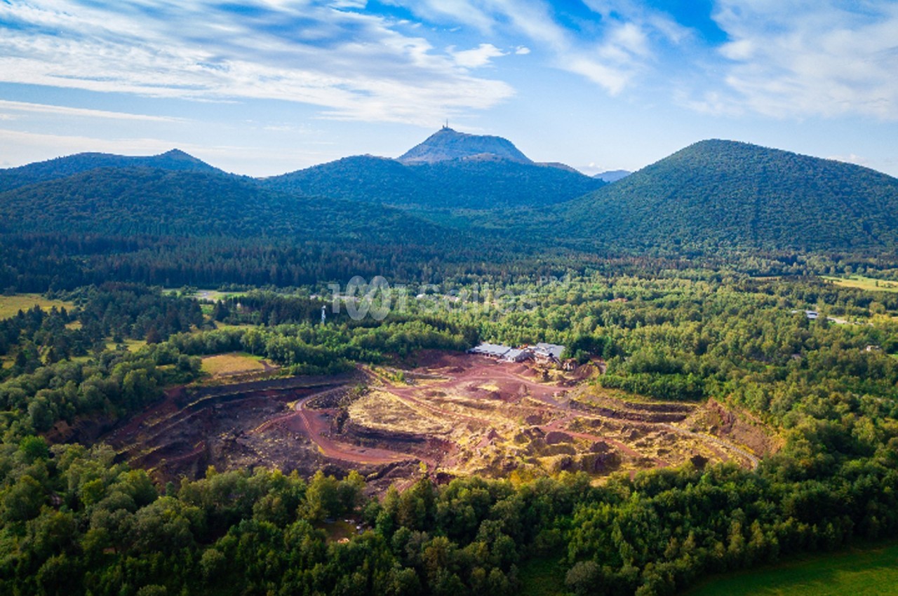 Vue aérienne d'une carrière entourée de forêts avec des montagnes en arrière-plan sous un ciel partiellement nuageux.