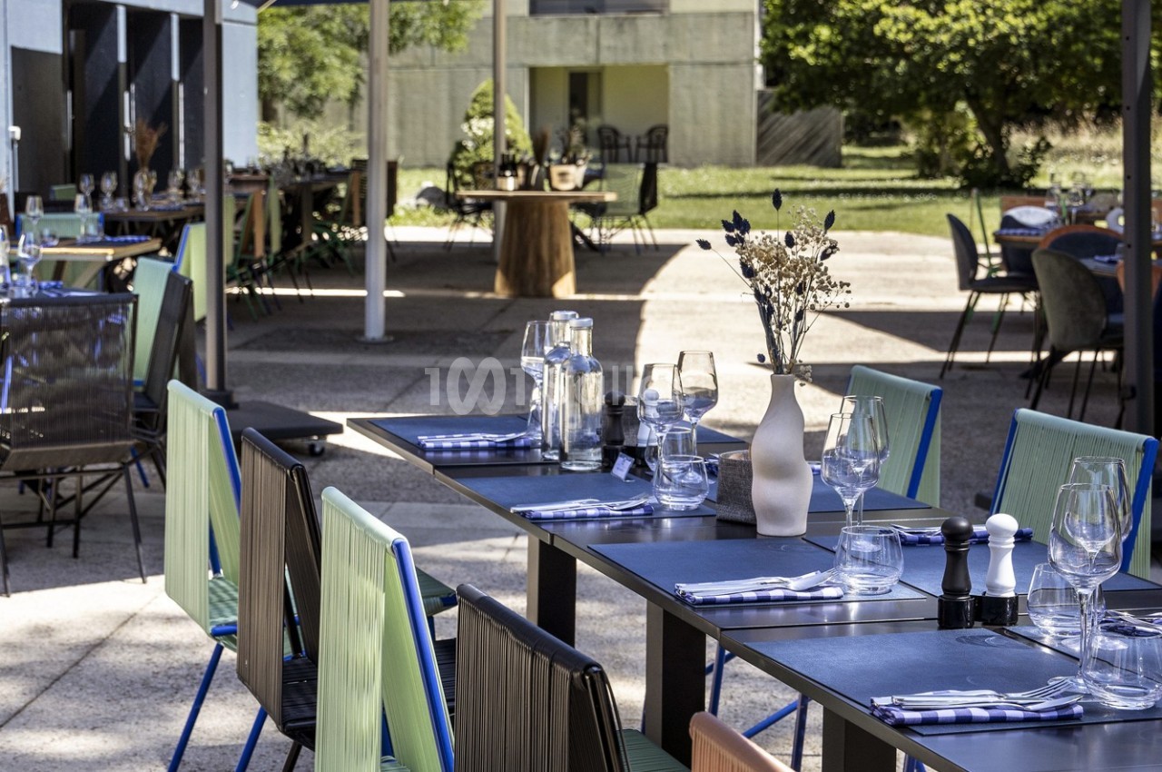 Terrasse d'un restaurant avec tables dressées, chaises colorées et vue sur un jardin en arrière-plan.