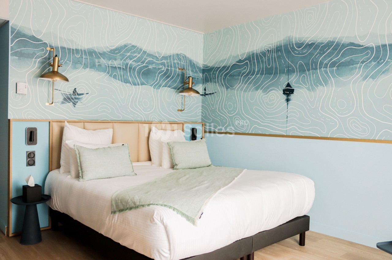 Lit double avec coussins verts dans une chambre lumineuse, décorée d'une fresque murale représentant un paysage abstrait.
