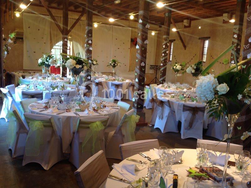 Salle décorée pour un événement avec tables rondes dressées, nappes blanches, fleurs et chaises ornées de rubans verts.